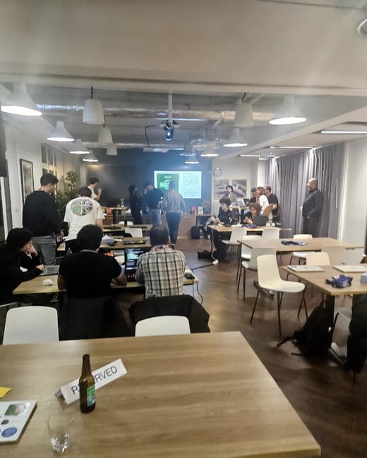 A candid photograph captures an indoor event, likely a workshop or hackathon, in a modern co-working space, showing approximately two dozen people working on laptops or gathered near a projected screen in the background. A table in the immediate foreground holds a beer bottle, a glass, and a sign reading 'P RVED'.