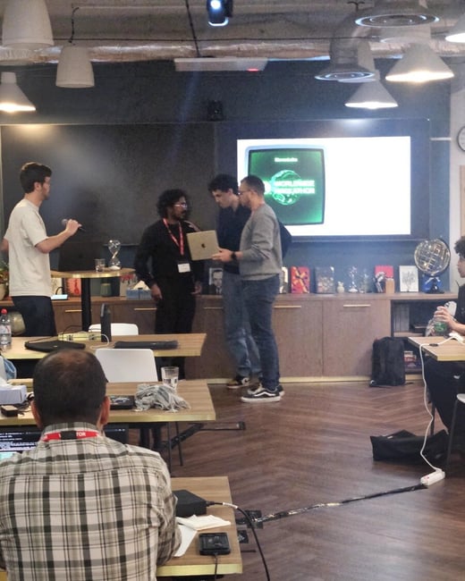 A candid photograph taken indoors at a technical event, likely a hackathon, showing several male participants standing and discussing a laptop near a large screen displaying the text "WORLDWIDE HACKATHON," while another participant is seated in the foreground working at a desk.