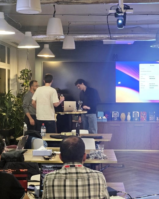 A candid photograph taken indoors at what appears to be a tech event or presentation, showing several people gathered near a large display screen in the background while attendees work on laptops at wooden tables in the foreground.