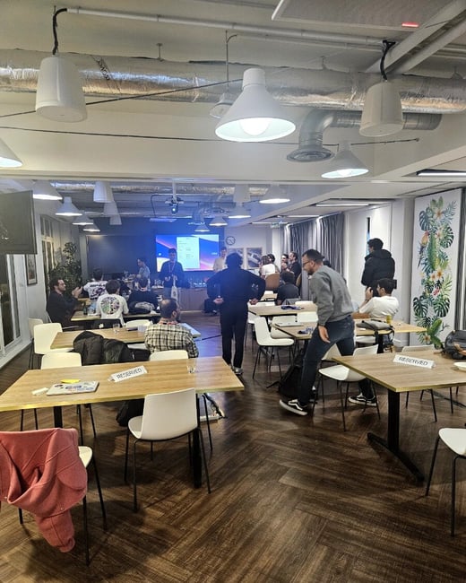 A candid photograph captures an indoor professional or social gathering taking place in a modern venue with exposed ceilings and wooden floors, where attendees are mingling and observing a presentation screen in the background.