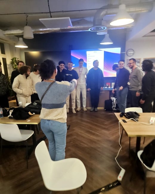 An indoor photograph capturing a social gathering where a person, seen from the back, appears to be taking a picture of a large group of colleagues posing in front of a brightly lit screen.