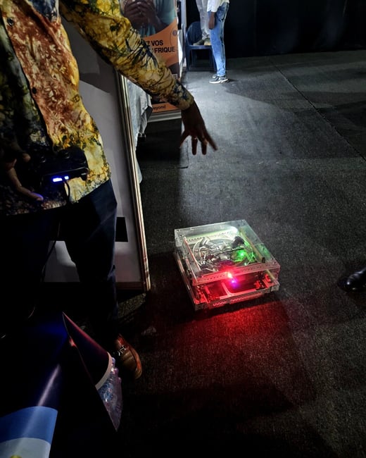 A person in a colorful patterned shirt is operating a small, transparent robot on the floor using a handheld controller at an indoor event.