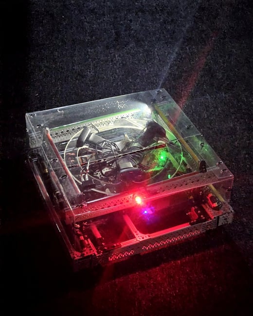 A small, box-shaped robot with a transparent acrylic top and visible internal wiring is illuminated by glowing red and green lights on a dark, textured surface.