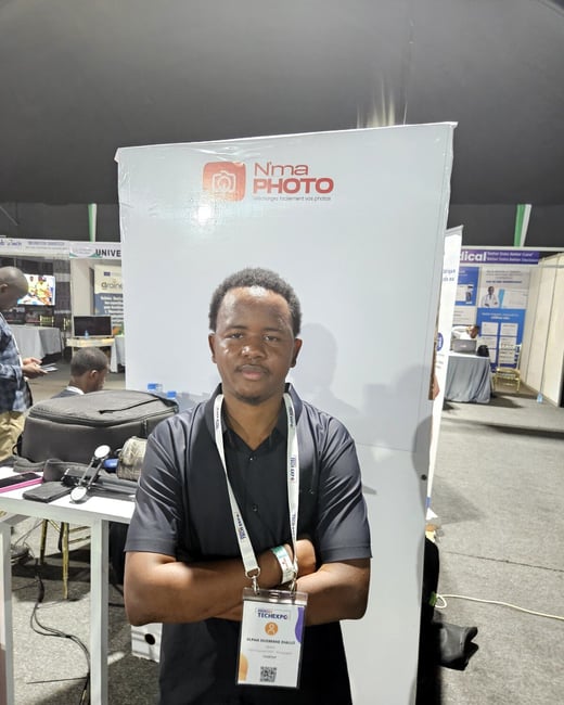 A man stands with his arms crossed in front of a white promotional booth at a tech event, wearing a lanyard and an identification badge.