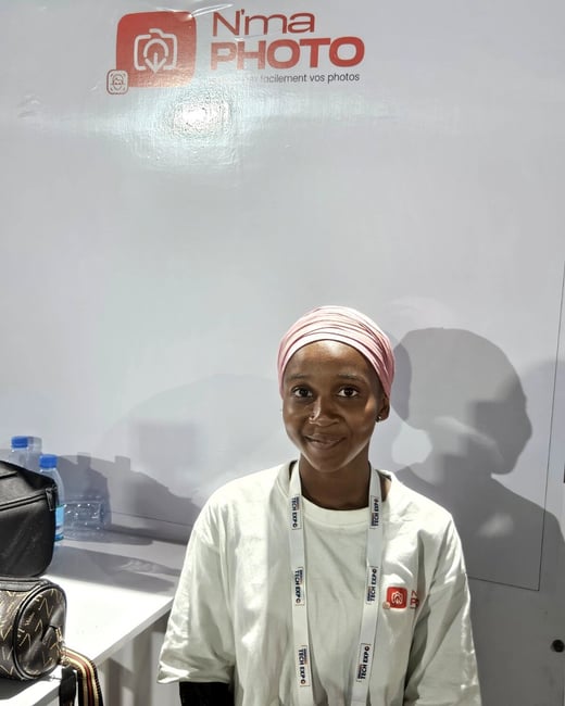 A woman wearing a pink headwrap and a white T-shirt with a lanyard stands in front of a white wall featuring a 'N'ma PHOTO' logo.