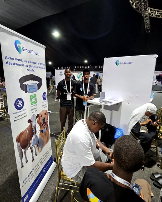 The image shows a promotional booth for SmarTrack, a company providing animal tracking solutions, at an indoor event. Two men in branded t-shirts stand behind a table, while others sit nearby.