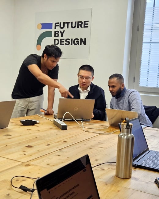 Three men are gathered around a wooden table in an office, looking at a laptop screen while one man points to something on it.