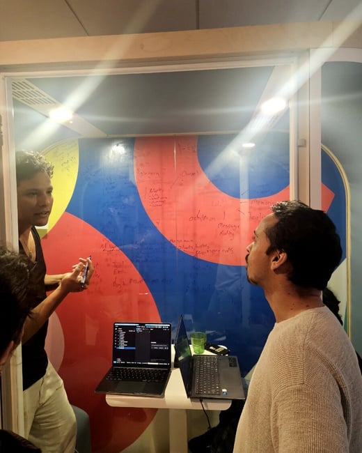 Two men are engaged in a discussion inside a glass-walled meeting pod, with one man gesturing towards a colorful whiteboard wall covered in handwritten notes.
