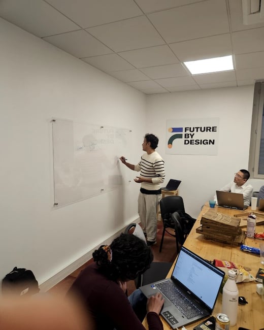 A man in a striped sweater writes on a glass whiteboard in an office setting while colleagues sit at a table with laptops and pizza boxes.