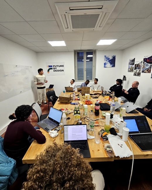 A group of people are gathered around a large wooden table in an office setting for a meeting or workshop. One person stands at a whiteboard while others sit with laptops, pizza boxes, and drinks.