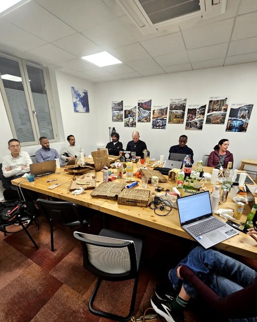A group of people are gathered around a large wooden table in an office setting, which is cluttered with pizza boxes, laptops, and various drinks.