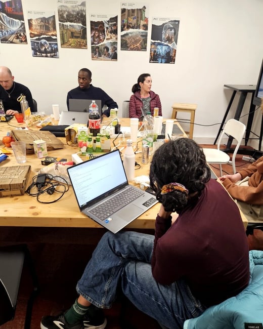 A group of people are gathered around a large wooden table in a workspace, using laptops and eating pizza. The room has posters on the wall and a casual, collaborative atmosphere.