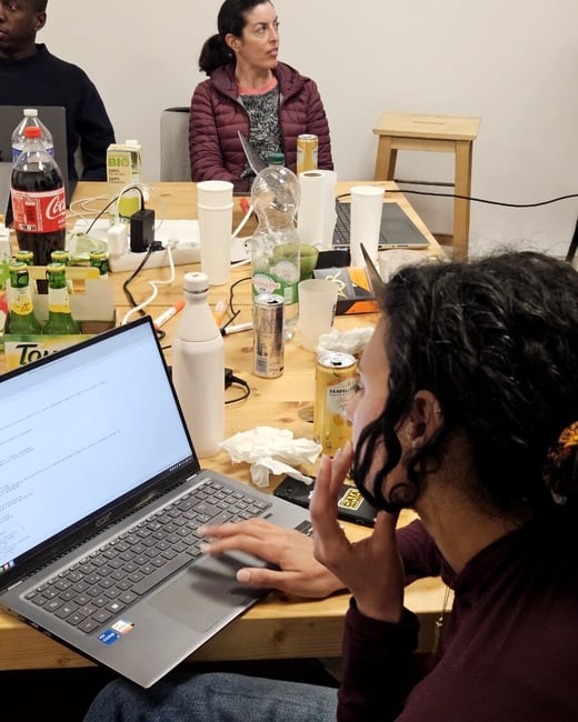 A group of people are gathered around a wooden table cluttered with drinks, snacks, and laptops, with one person in the foreground actively typing on a laptop.