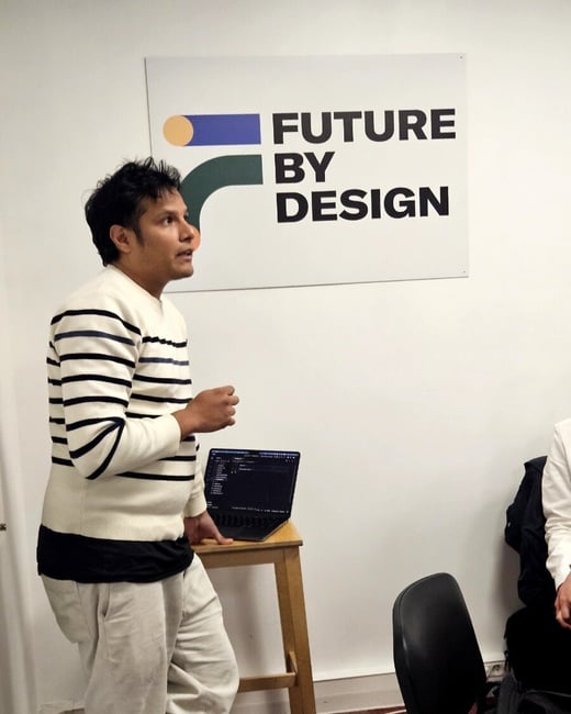 A man in a striped sweater stands in profile, gesturing while speaking in front of a 'FUTURE BY DESIGN' sign. A laptop displaying code sits on a wooden stool next to him.