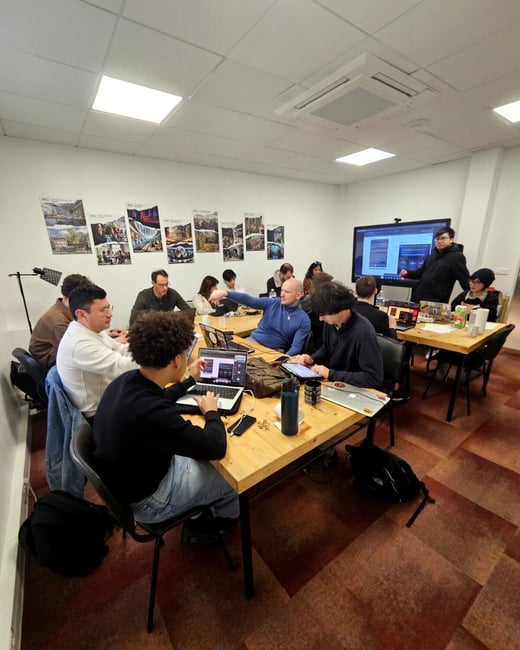 A group of people are gathered in a brightly lit room for a workshop or presentation, sitting at wooden tables with laptops while one person stands near a large screen.