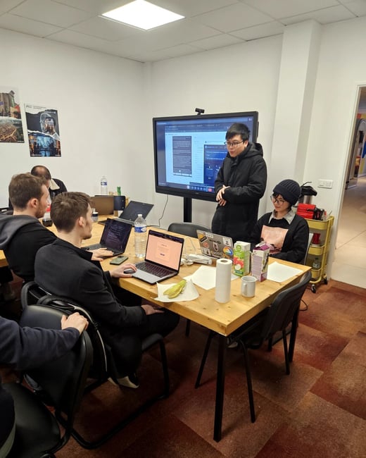 A group of people are gathered in an office setting for a presentation or collaborative session, with one person standing by a large screen while others sit at a table with laptops.