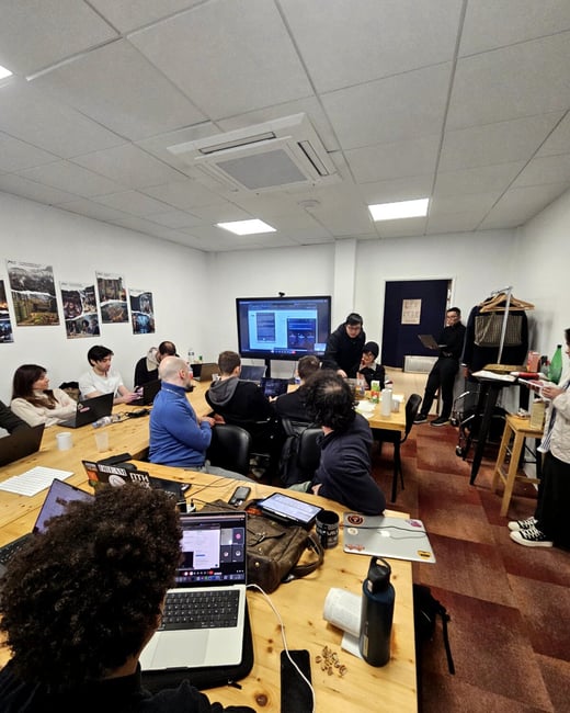 A group of people are gathered in an office or classroom setting, focused on a presentation shown on a large screen and their individual laptops.