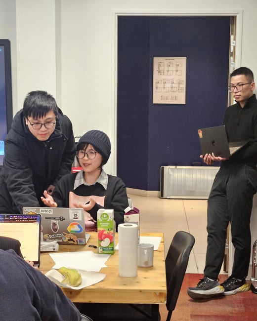 A group of people are gathered in an office setting, engaged in a discussion around a laptop placed on a wooden table.