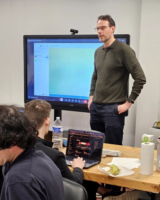 A man in a green sweater stands in front of a large digital screen during a presentation, while others sit at a wooden table with a laptop in the foreground.