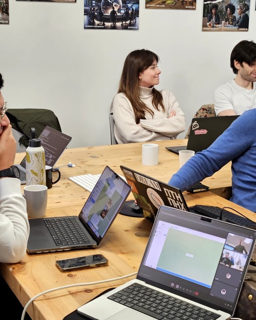 A group of people are working around a large wooden table in a modern office or coworking space, with several open laptops and a video call in progress.
