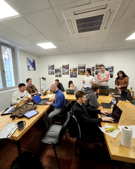 A group of people are gathered around a large wooden table in a bright office space, most of them working on laptops or engaged in a collaborative discussion.