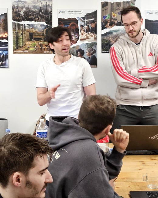 A man in a white t-shirt stands and gestures while speaking to a group in an office or workshop setting, with several posters on the wall behind him.