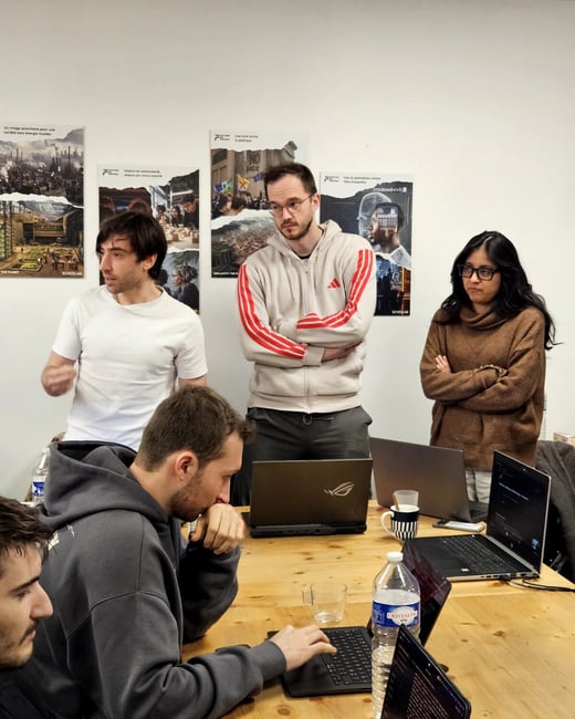 A group of young adults are gathered in a brightly lit room, some standing and some seated at a wooden table with laptops, appearing to be in a collaborative work or discussion session.