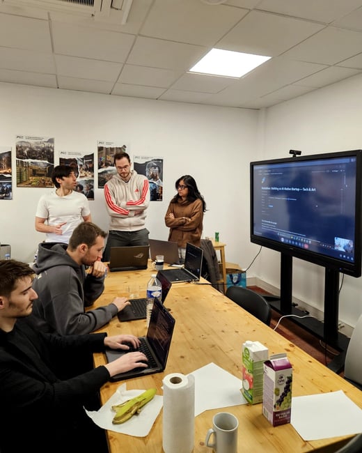 A group of five people are in a room, with some sitting at a long wooden table working on laptops and others standing and looking at a large digital screen displaying a hackathon project.
