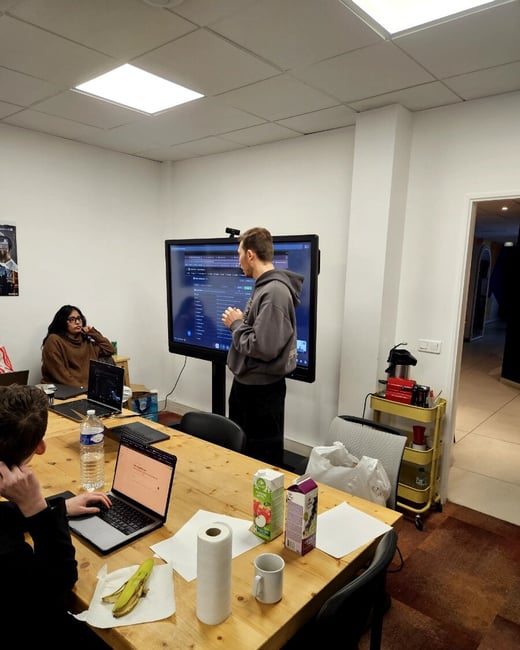 A man stands in an office giving a presentation or demonstration on a large screen to a small group seated at a wooden table with laptops and snacks.