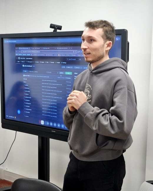 A young man in a grey hoodie stands in front of a large digital screen displaying a GitHub repository, likely giving a project presentation.