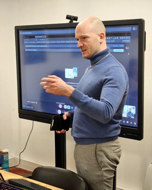 A man in a blue turtleneck sweater is giving a presentation in front of a large interactive display showing a Google Meet session.