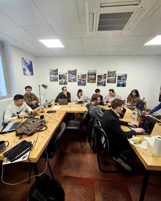 A group of people are gathered around a large wooden table in a brightly lit room, most of them focused on their laptops.