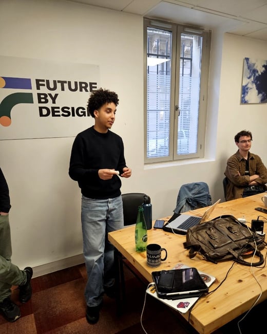 A young man with curly hair stands and speaks in an office setting, while another man sits at a wooden table in the background.