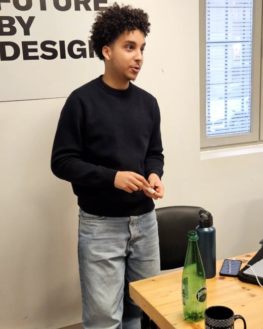 A young man with curly hair, wearing a black sweater and light-blue jeans, stands in an indoor setting and appears to be speaking or giving a presentation.