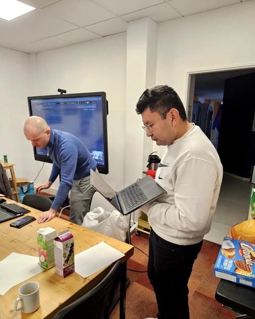 Two men are in an office or meeting room; one stands while looking at a laptop he is holding, and the other leans over a wooden table near a large screen.