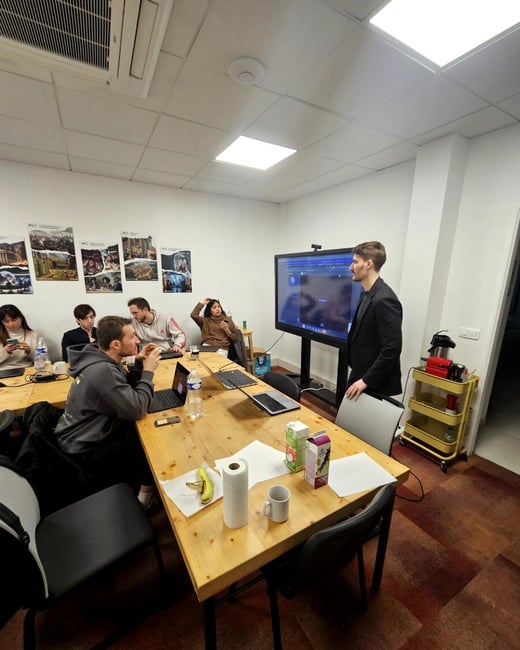 A group of people are gathered in a meeting room for a presentation or demo, with one person standing next to a large screen and others seated at a wooden table with laptops.