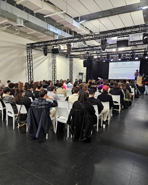 A large audience is seated in an industrial-style hall, watching a presentation on a stage equipped with a large screen and professional lighting rigs.
