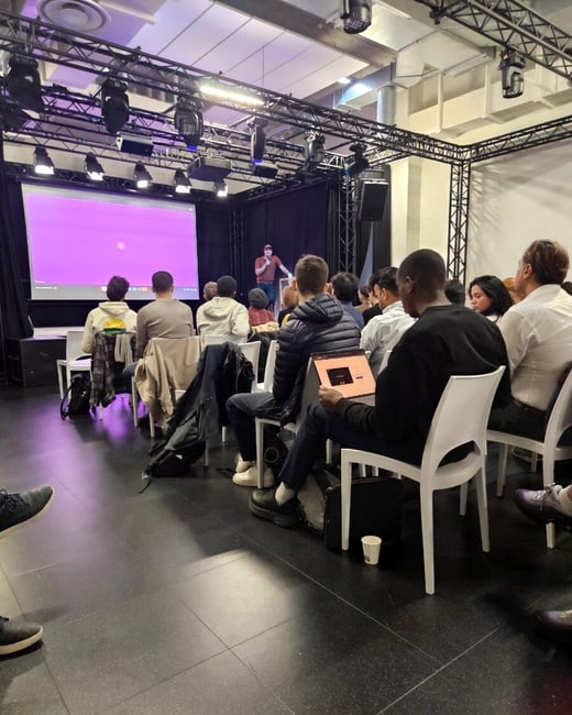 A photograph of a presentation event where an audience is seated in a dark room, watching a speaker on a small stage next to a large purple screen.