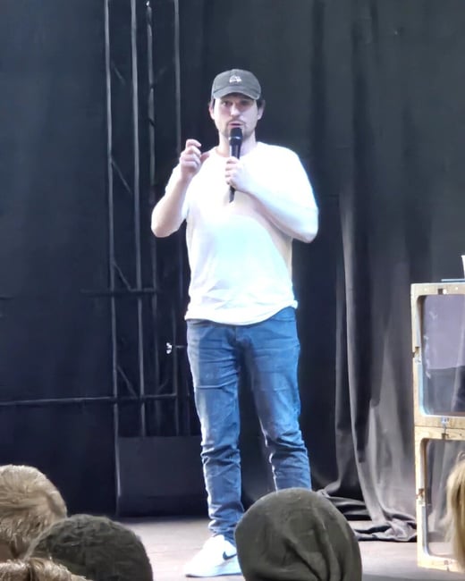 A man in a white t-shirt and jeans stands on a stage, speaking into a microphone in front of an audience.