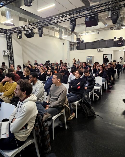 A wide-angle photograph showing a large audience of young adults seated in rows of white chairs within an industrial-style event space, likely attending a talk or presentation.