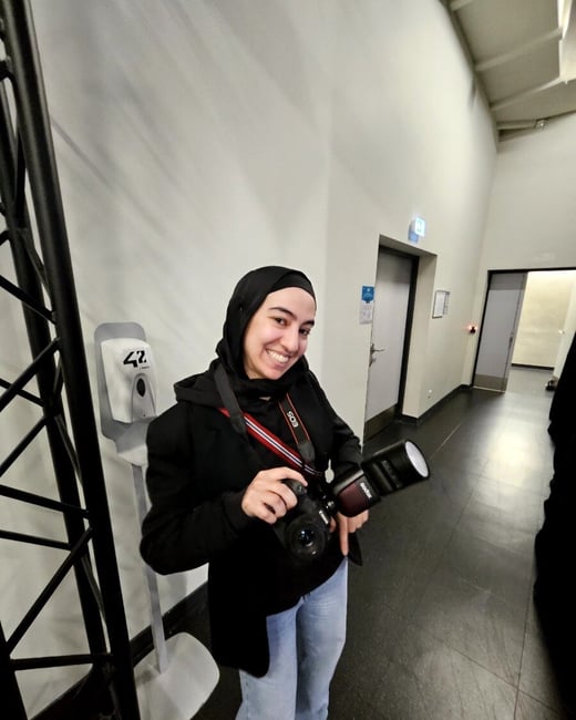 A smiling woman wearing a black hijab and jacket holds a professional camera with an external flash in an indoor hallway.