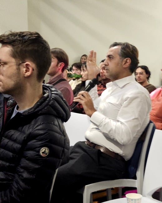 A man in a white shirt sits in an audience, holding a microphone and raising his hand to speak during a presentation or event.