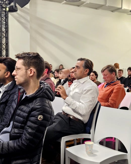 An indoor scene showing an audience seated in white chairs, with a man in a white shirt holding a microphone as if preparing to ask a question.