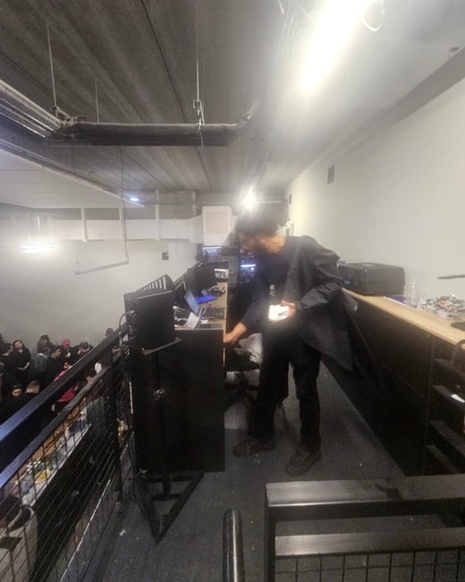 A person stands at a control desk on a mezzanine level, overlooking a crowd of people in a large indoor venue.
