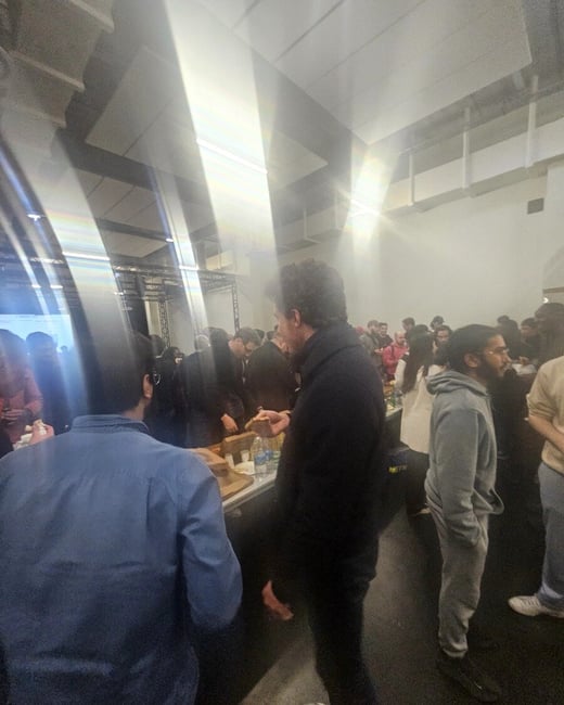 A crowded indoor social gathering where people are standing around tables with food and drinks, featuring a prominent lens flare on the left side.