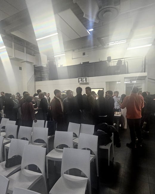 A social gathering or event inside a large hall with rows of white chairs in the foreground and a crowd of people standing and talking in the background.
