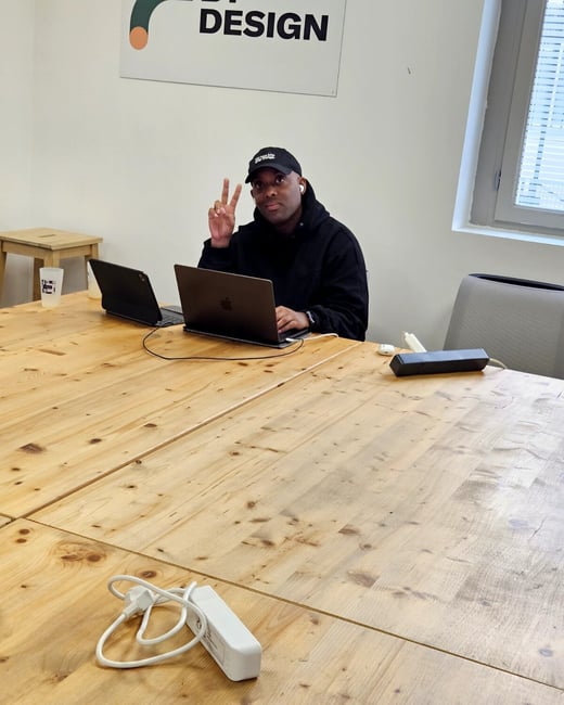 A man in a black hoodie and cap is sitting at a large wooden table with a laptop, smiling and making a peace sign for the camera.