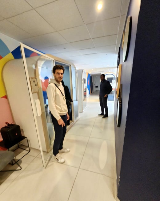 A modern office hallway featuring two men standing near glass-walled privacy booths with colorful geometric wall art.