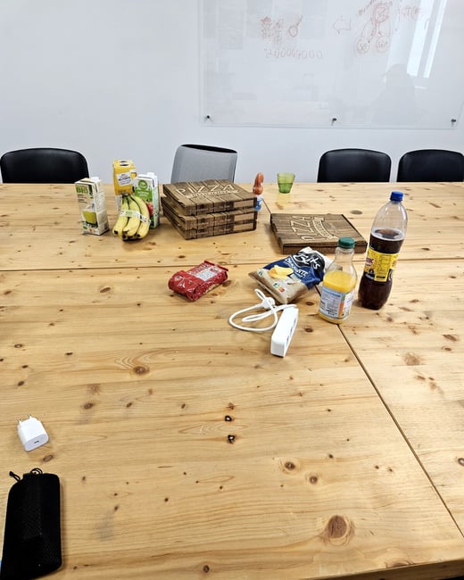 A wooden conference table is covered with various food items, including pizza boxes, chips, fruit, and drinks, in front of a whiteboard.