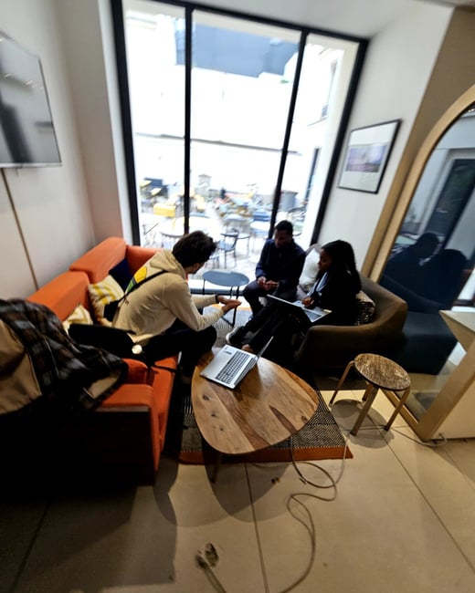 Three people are sitting in a modern lounge area, engaged in a collaborative discussion with laptops around a wooden coffee table.